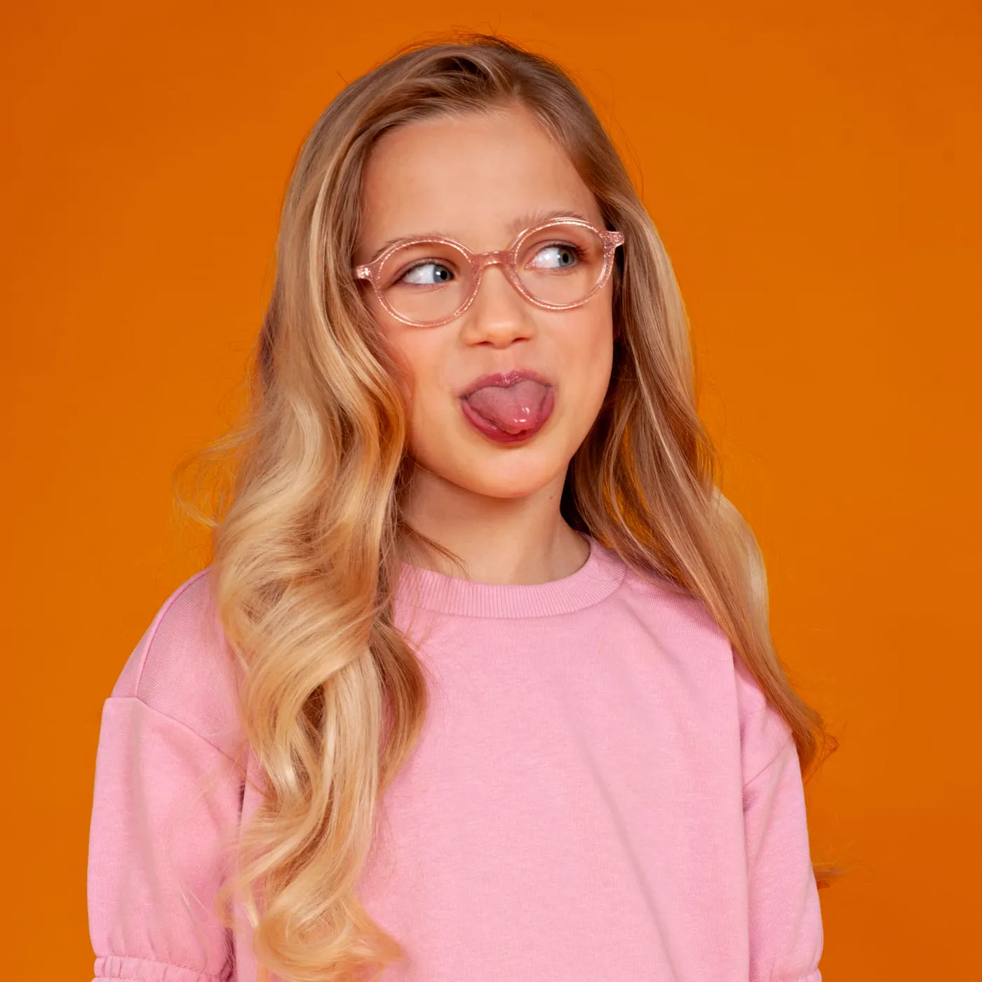 Meisje met een kinderbril van BBIG steekt haar tong uit, voor een oranje achtergrond